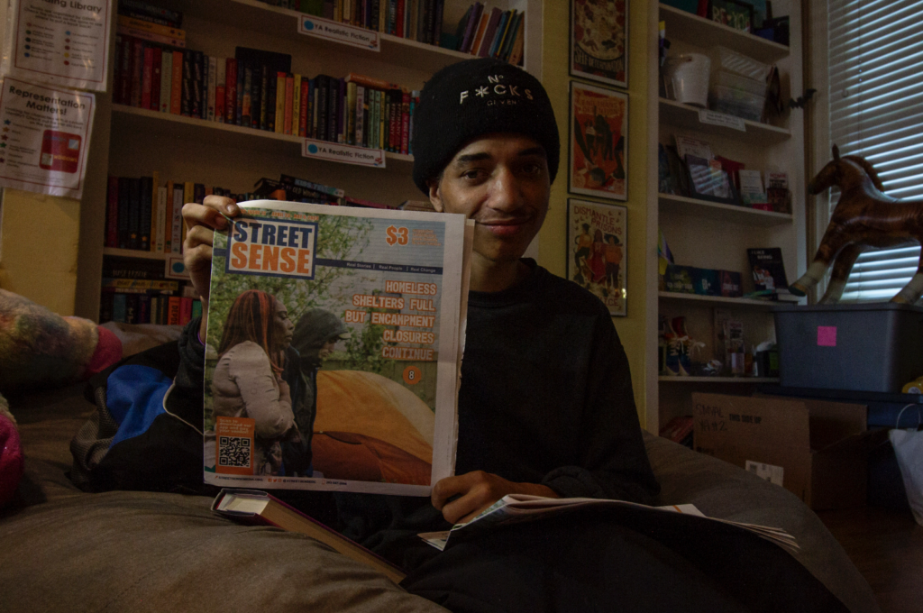 A youth holds up a copy of a newspaper he is featured on in front of a yellow bookcase.